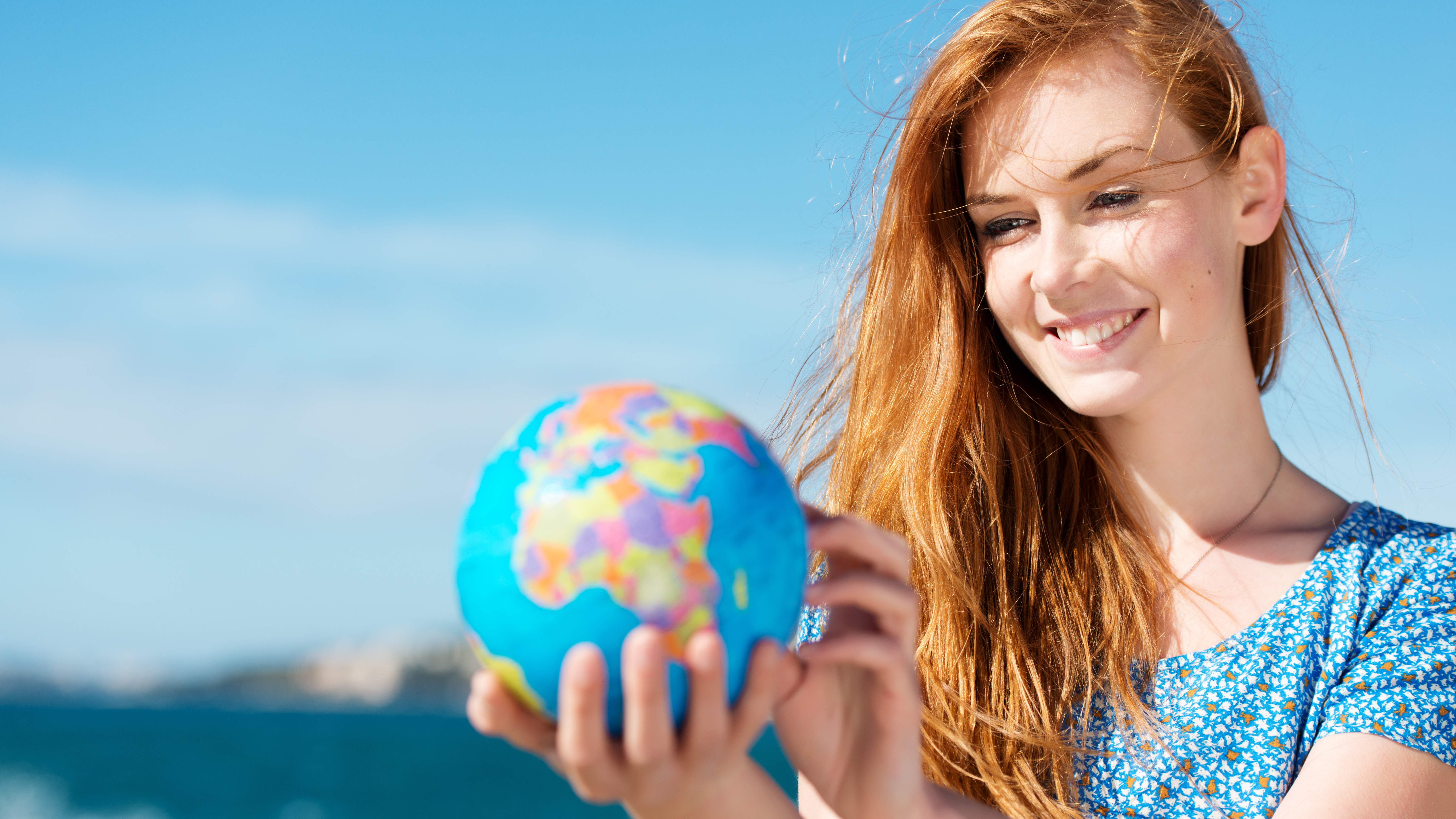 Eine Frau hält eine Weltkugel in der Hand und schaut lächelnd auf diese. Im Hintergrund sieht man das Meer.